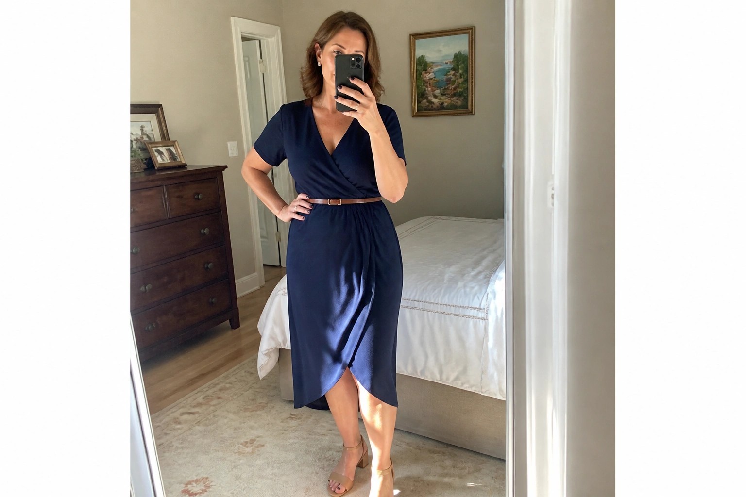 Woman in navy blue wrap dress with brown belt taking mirror selfie in bedroom.