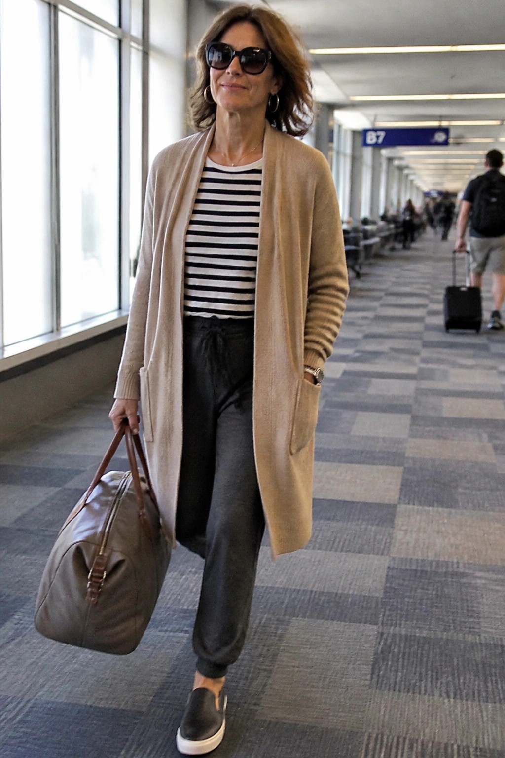 Woman in beige cardigan over striped top carrying leather bag in airport hallway