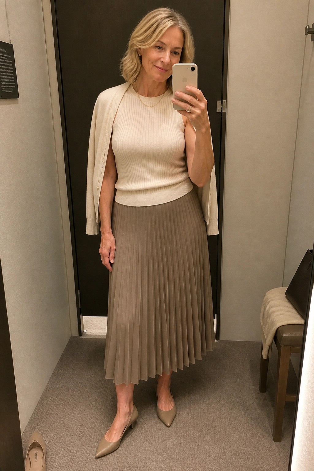 Woman in cream ribbed top and taupe pleated skirt taking mirror selfie.