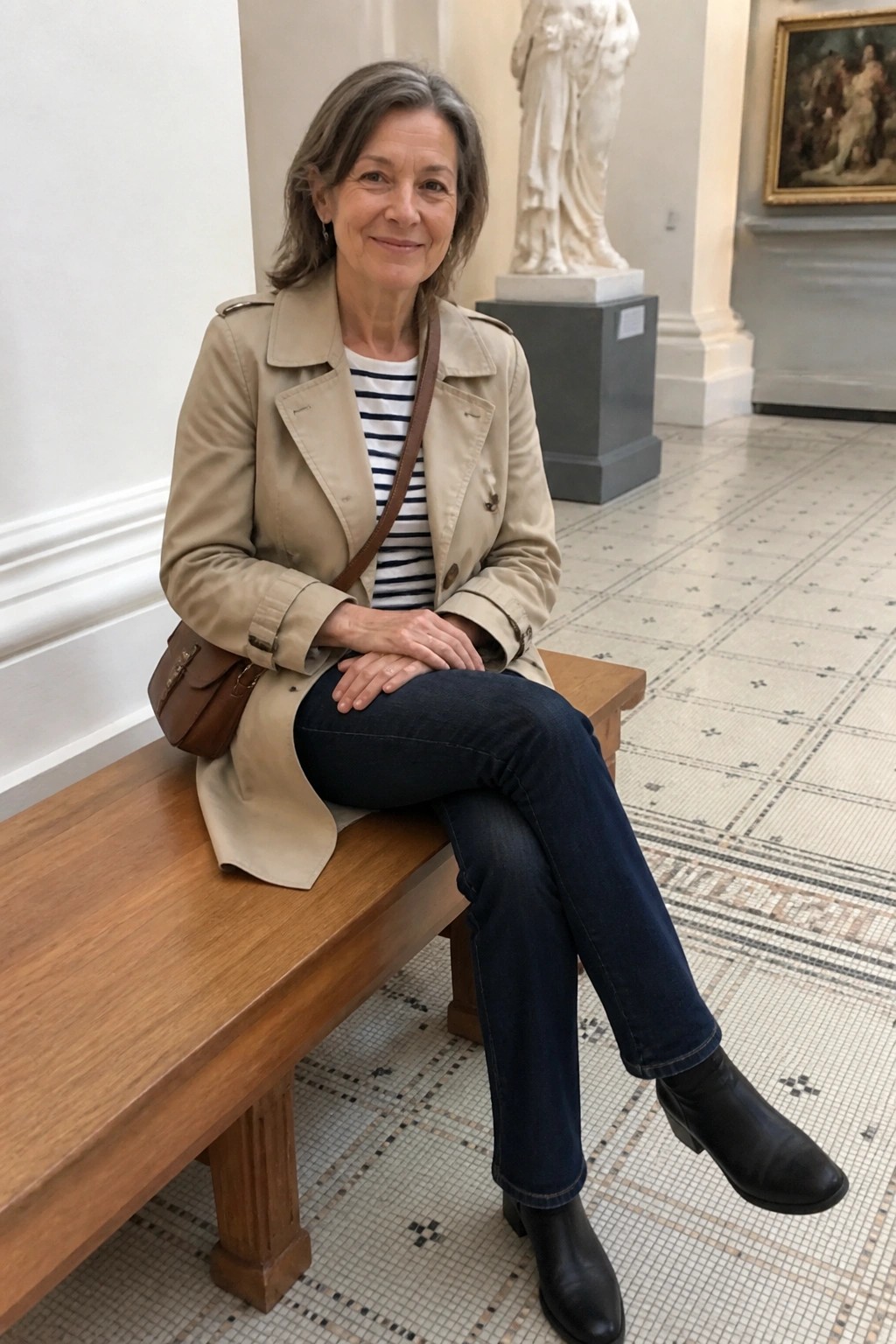 Woman in beige trench coat and jeans seated on bench in museum gallery