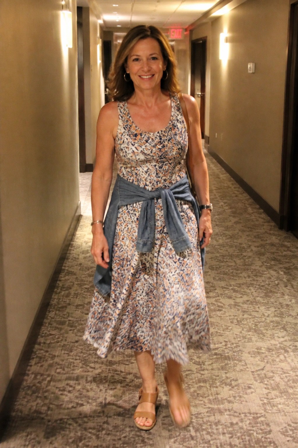 Smiling woman in floral dress with denim jacket tied at waist in hallway