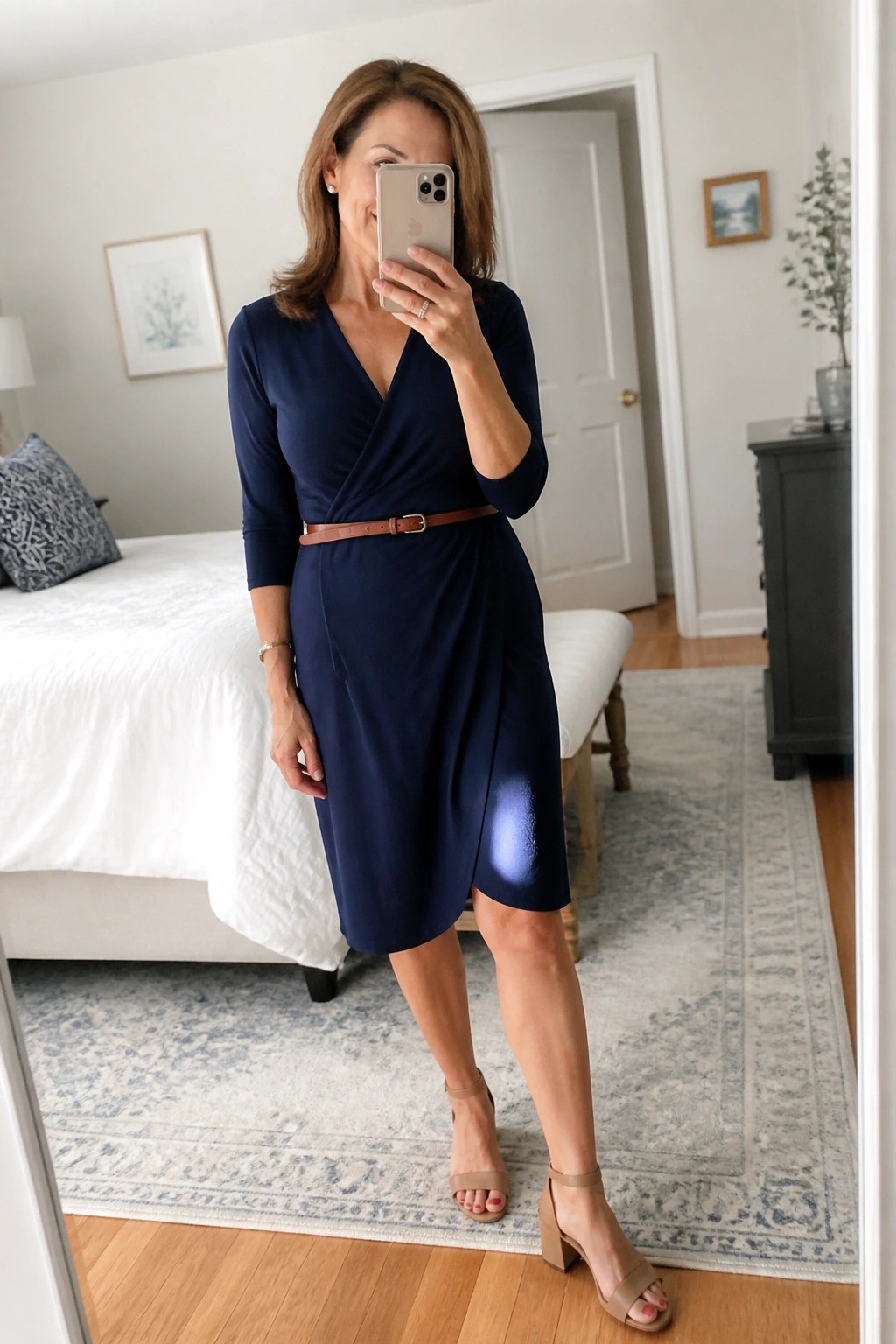 Woman in navy blue wrap dress with brown belt taking mirror selfie in bedroom.