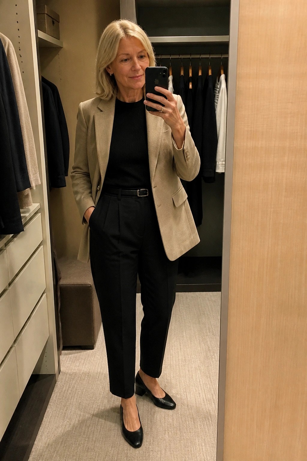 Blonde woman in beige blazer and black pants taking mirror selfie in closet