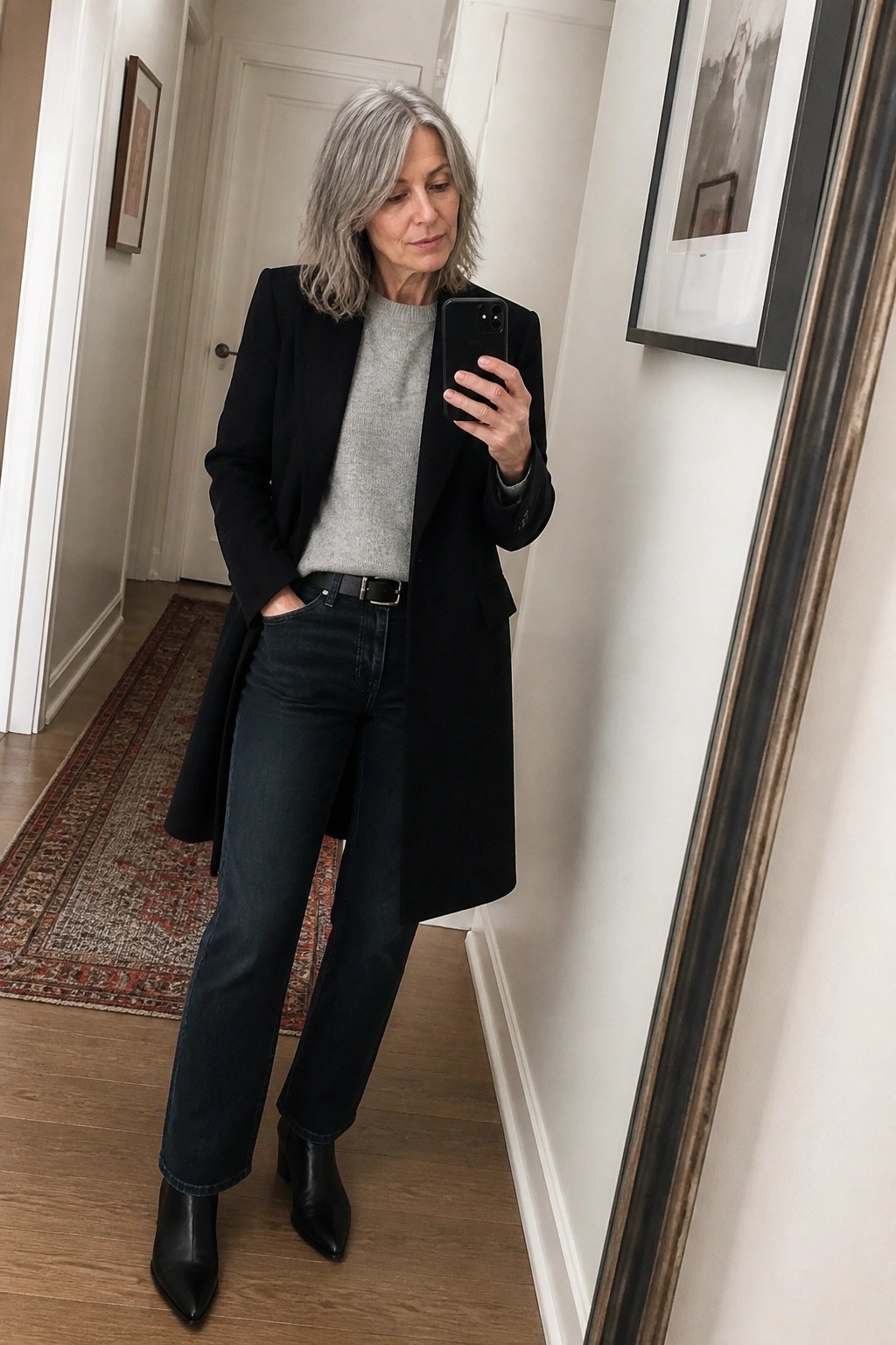 Gray-haired woman in black coat, gray sweater, and jeans taking mirror selfie