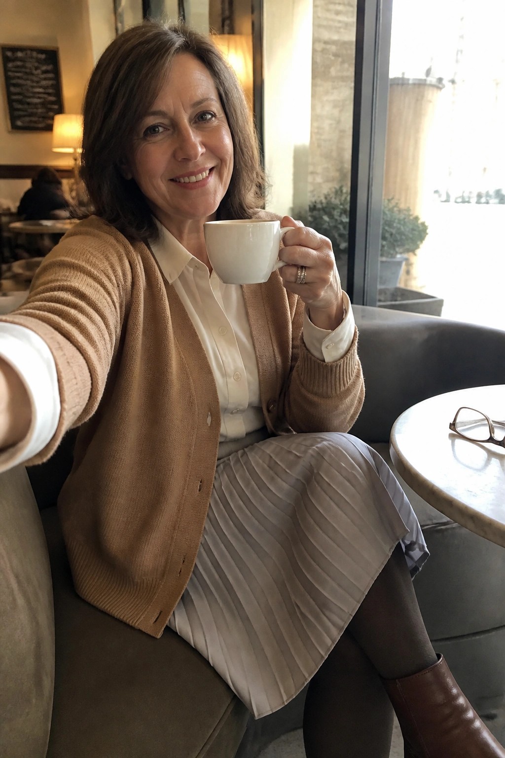 Smiling woman in brown cardigan and pleated skirt holding white coffee cup in cafe