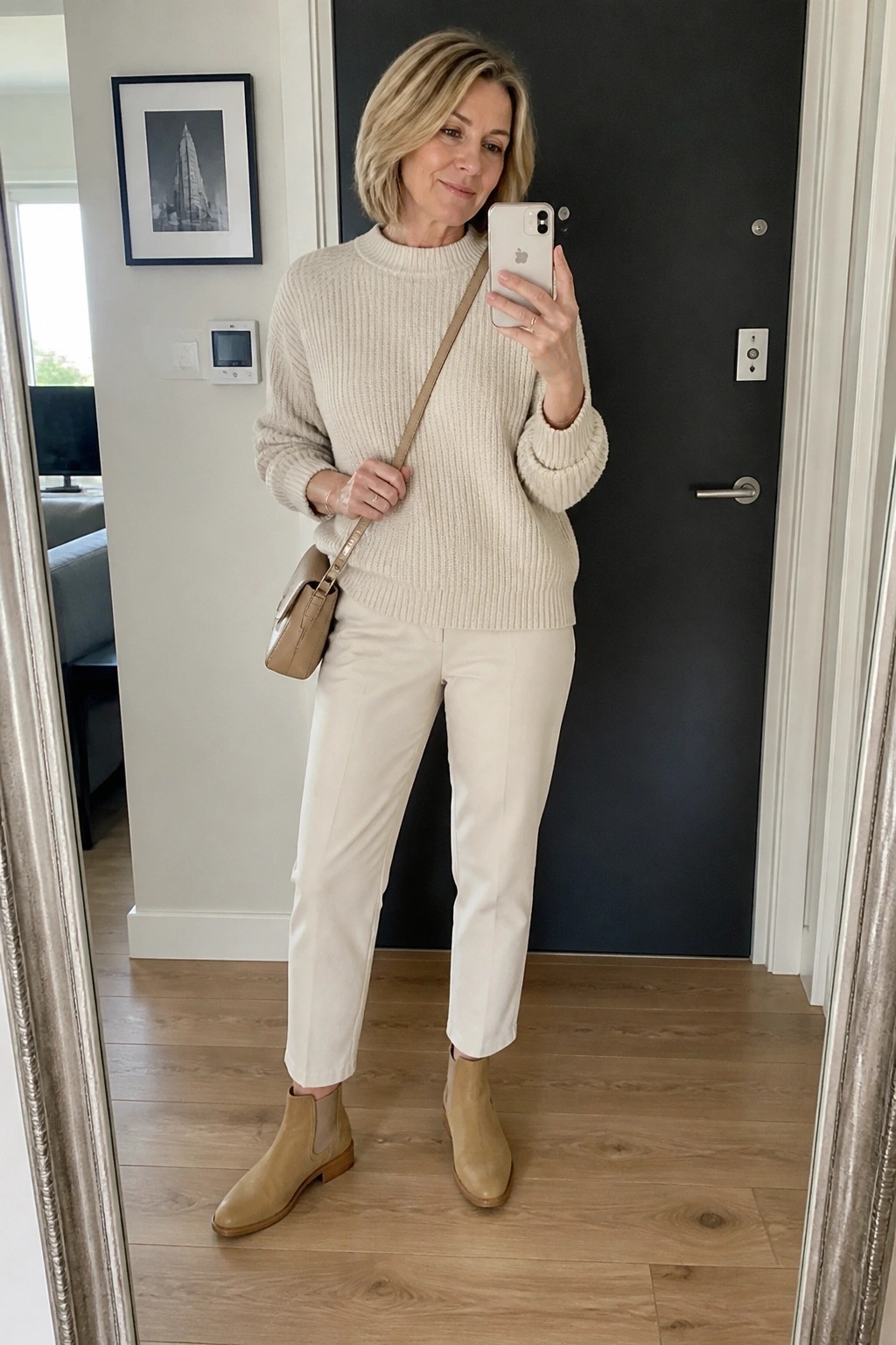 Woman in cream sweater and white pants taking mirror selfie with phone