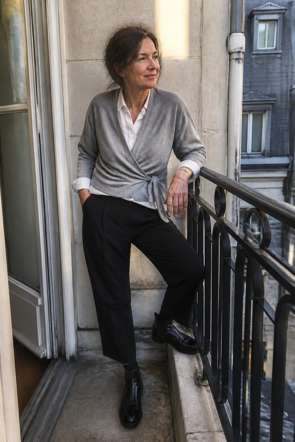 Woman in gray wrap sweater and black pants leaning on balcony railing