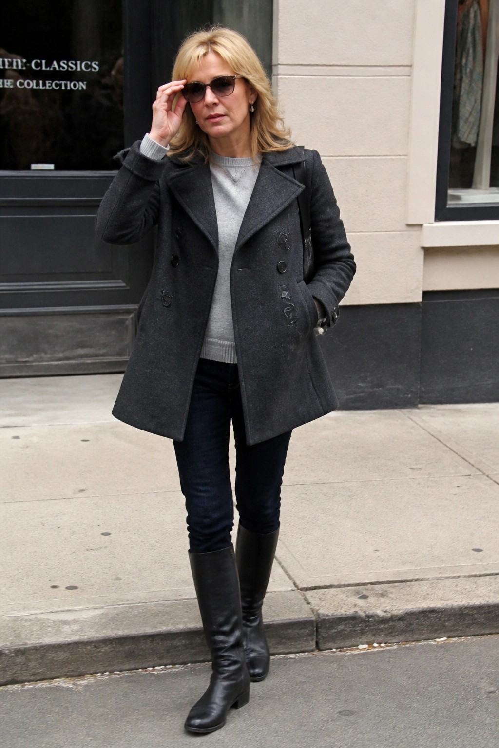 Blonde woman in gray coat and black boots adjusts sunglasses on sidewalk