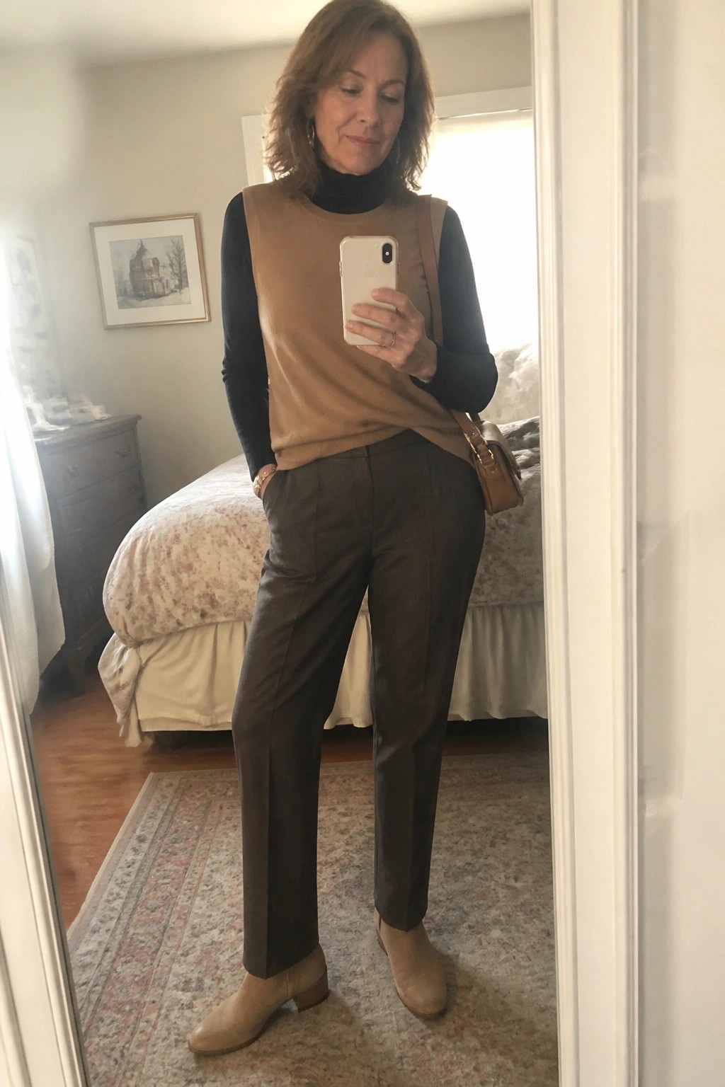 Auburn-haired woman in tan vest over black turtleneck and brown pants in bedroom mirror