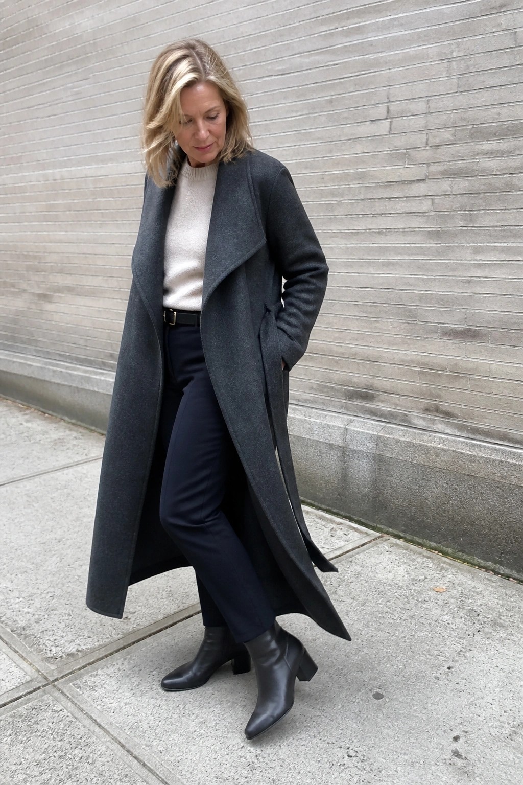 Woman in long gray coat over beige sweater beside brick wall on sidewalk