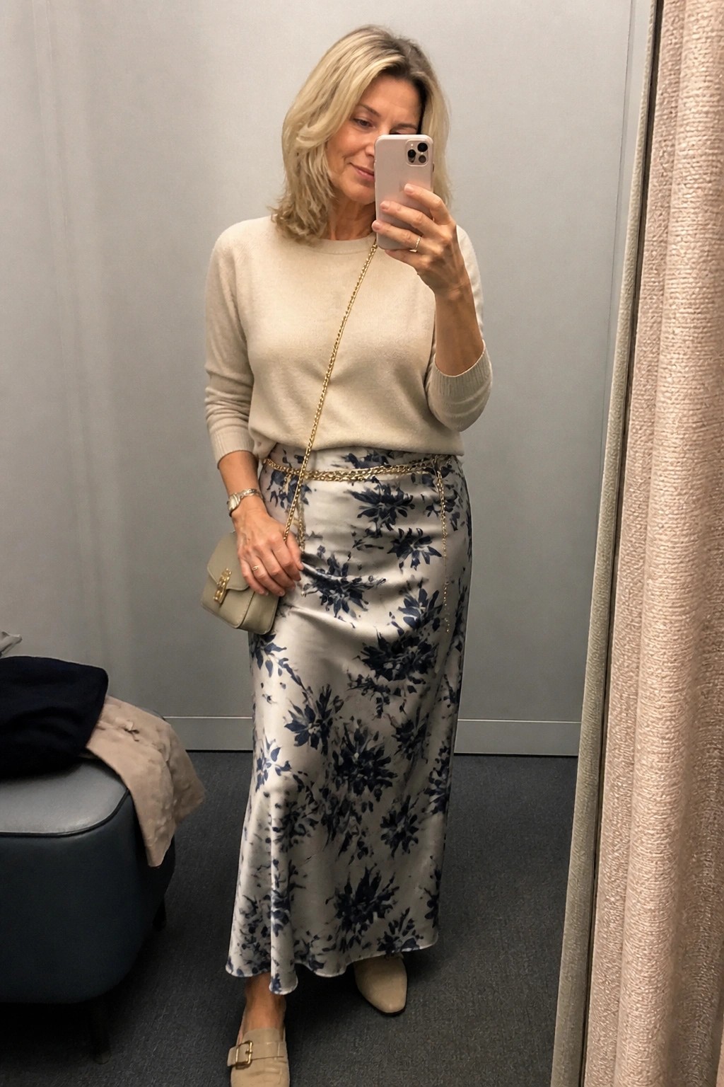 Blonde woman in cream sweater and blue floral satin skirt taking mirror selfie
