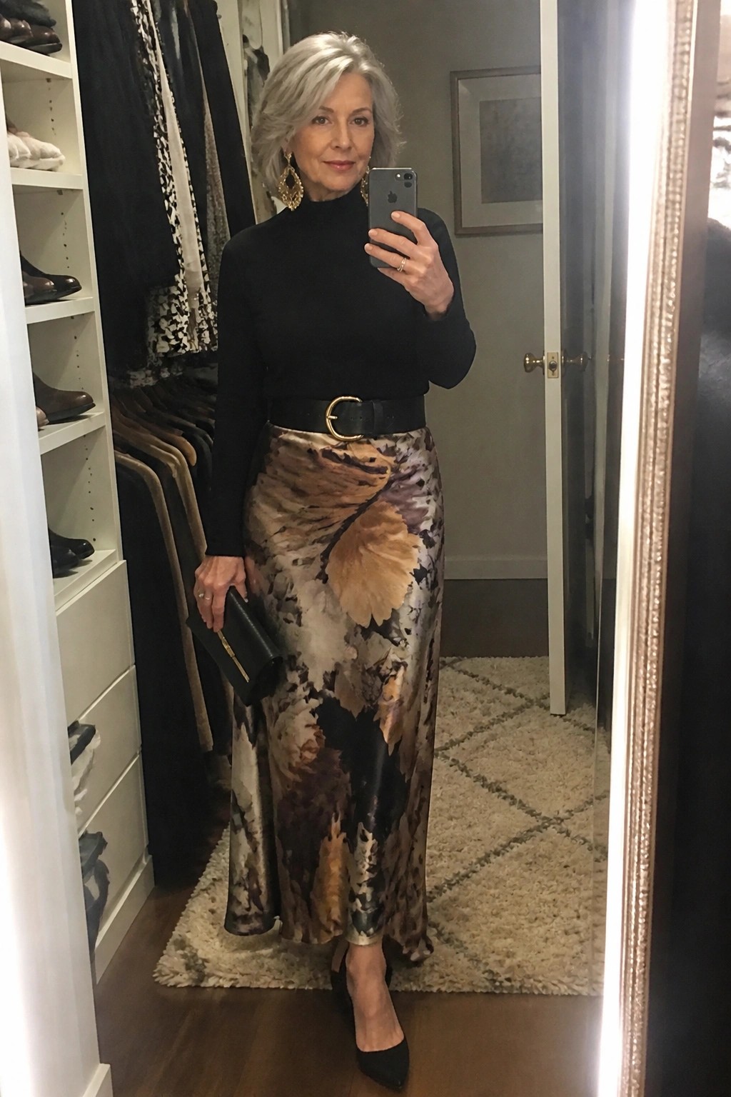 Gray-haired woman in black turtleneck and abstract floral skirt taking mirror selfie in closet