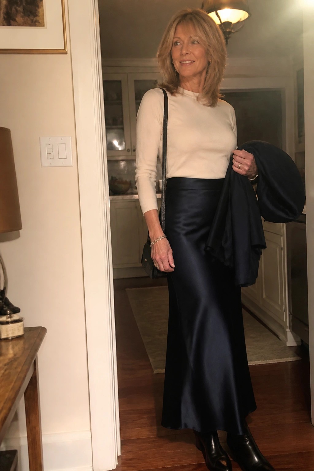 Woman in cream sweater and long navy satin skirt holding jacket indoors