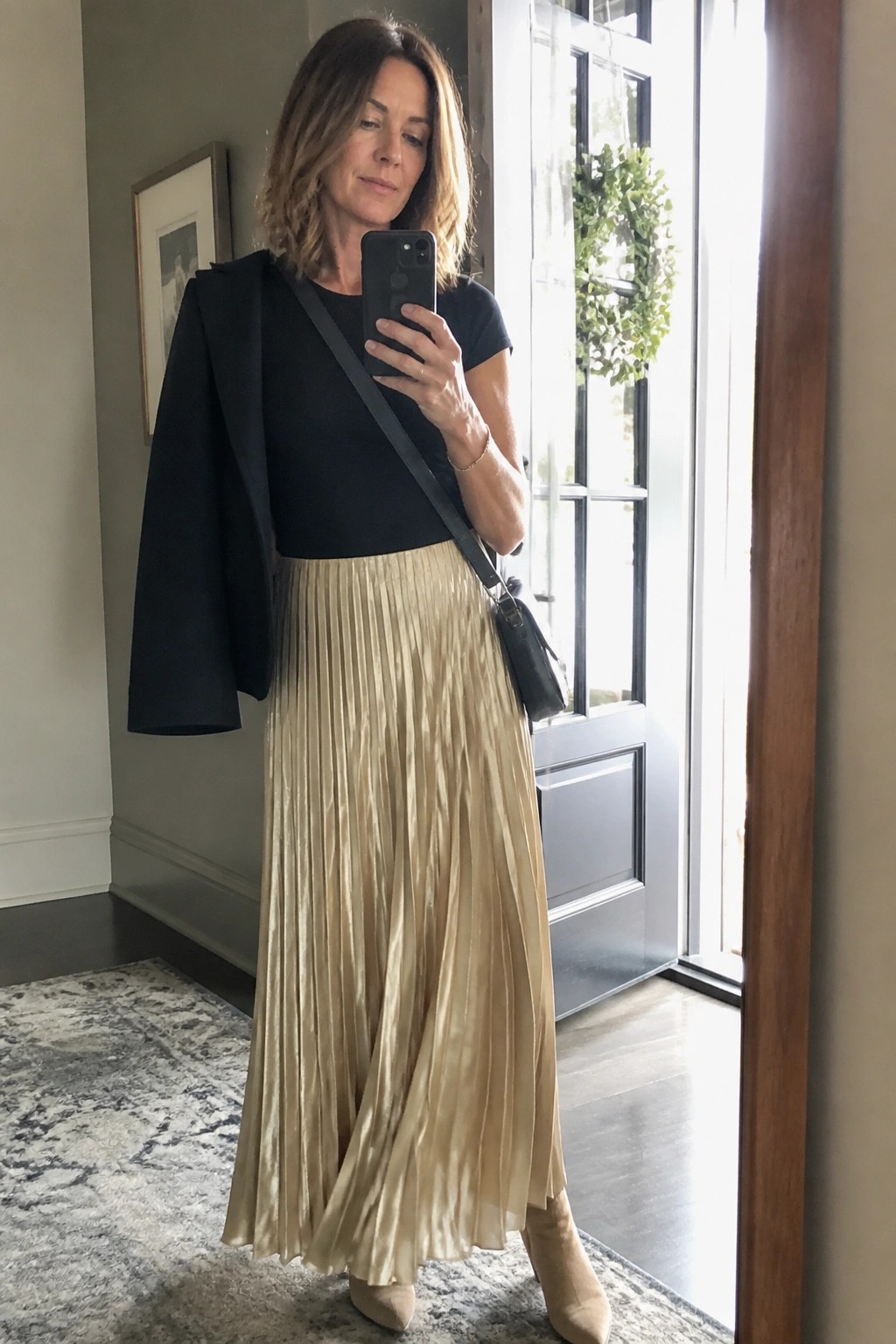 Woman over 50 in a black short sleeve top with black blazer draped over shoulders, gold satin pleated maxi skirt, beige ankle boots and black crossbody bag taking a mirror selfie.