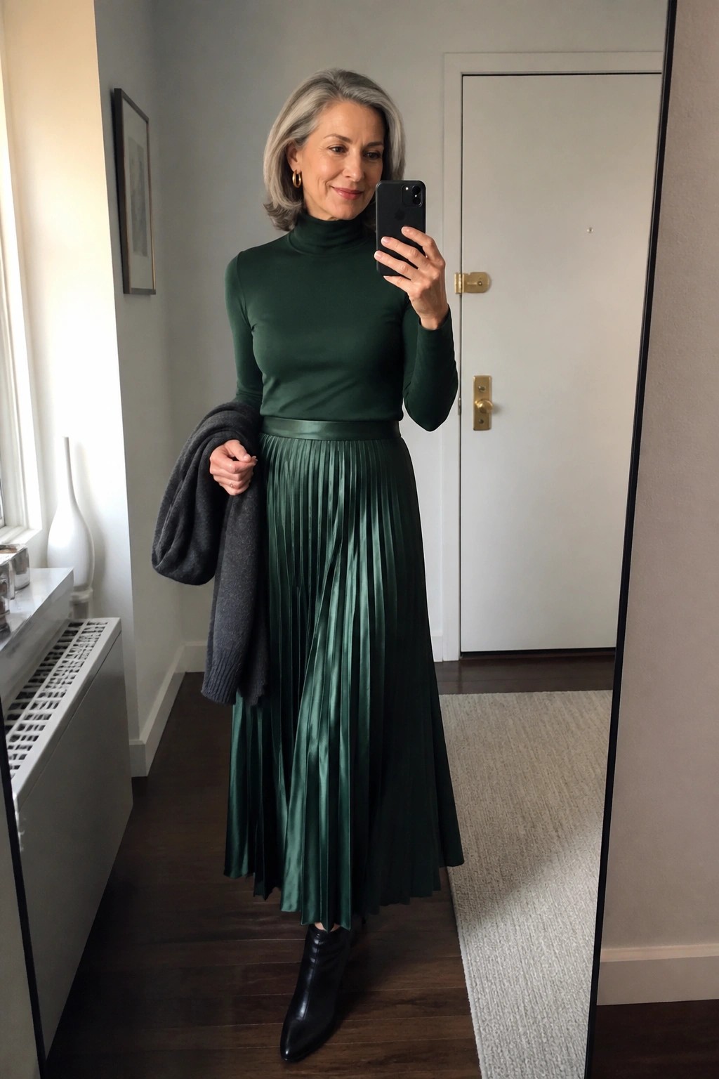 Woman with gray hair in green turtleneck and pleated skirt taking mirror selfie