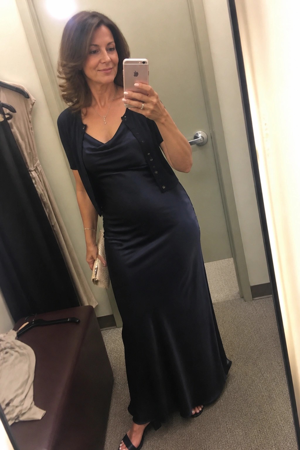 Woman in long navy satin dress and black cardigan taking mirror selfie with phone.