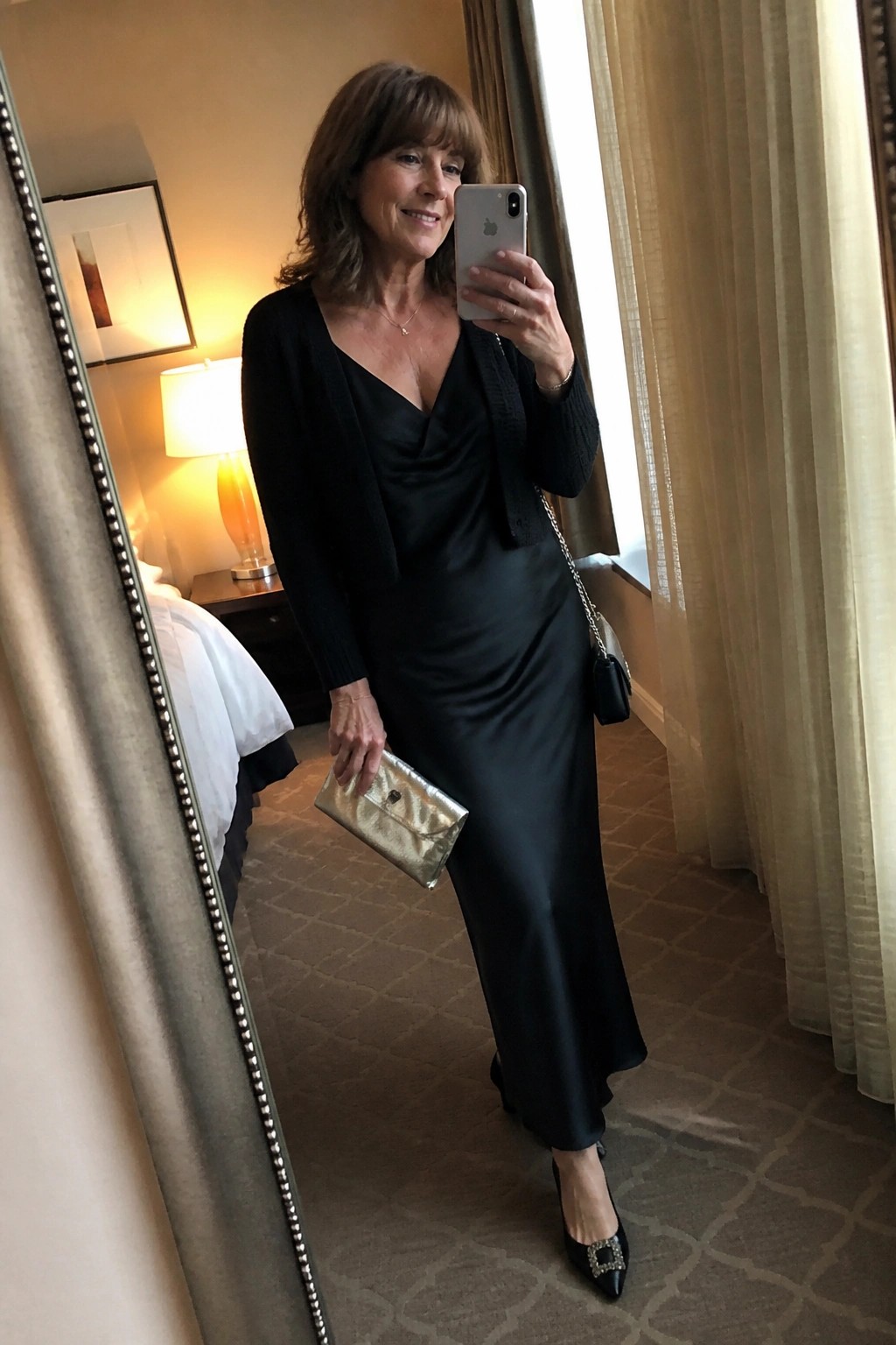 Woman over 50 in black satin maxi dress layered with black knit cardigan paired with black embellished heels and gold clutch while holding a phone.