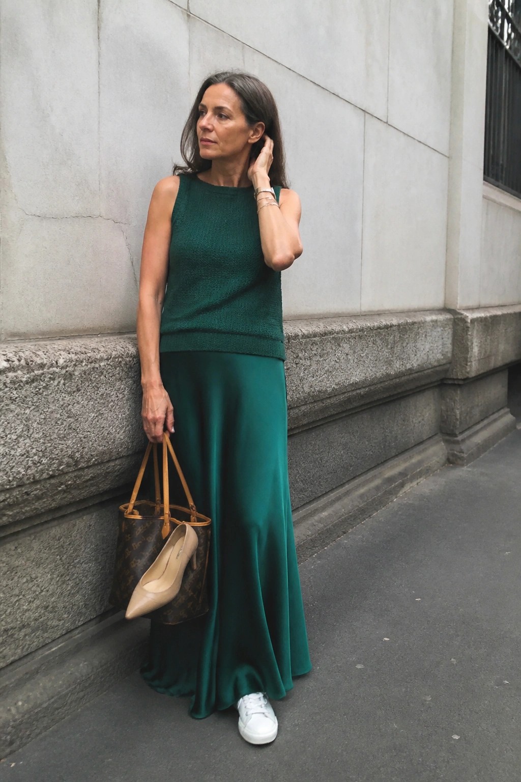 Woman in emerald green satin maxi skirt and matching sleeveless knit top with white sneakers and stacked bracelets holding a monogram tote containing beige heels