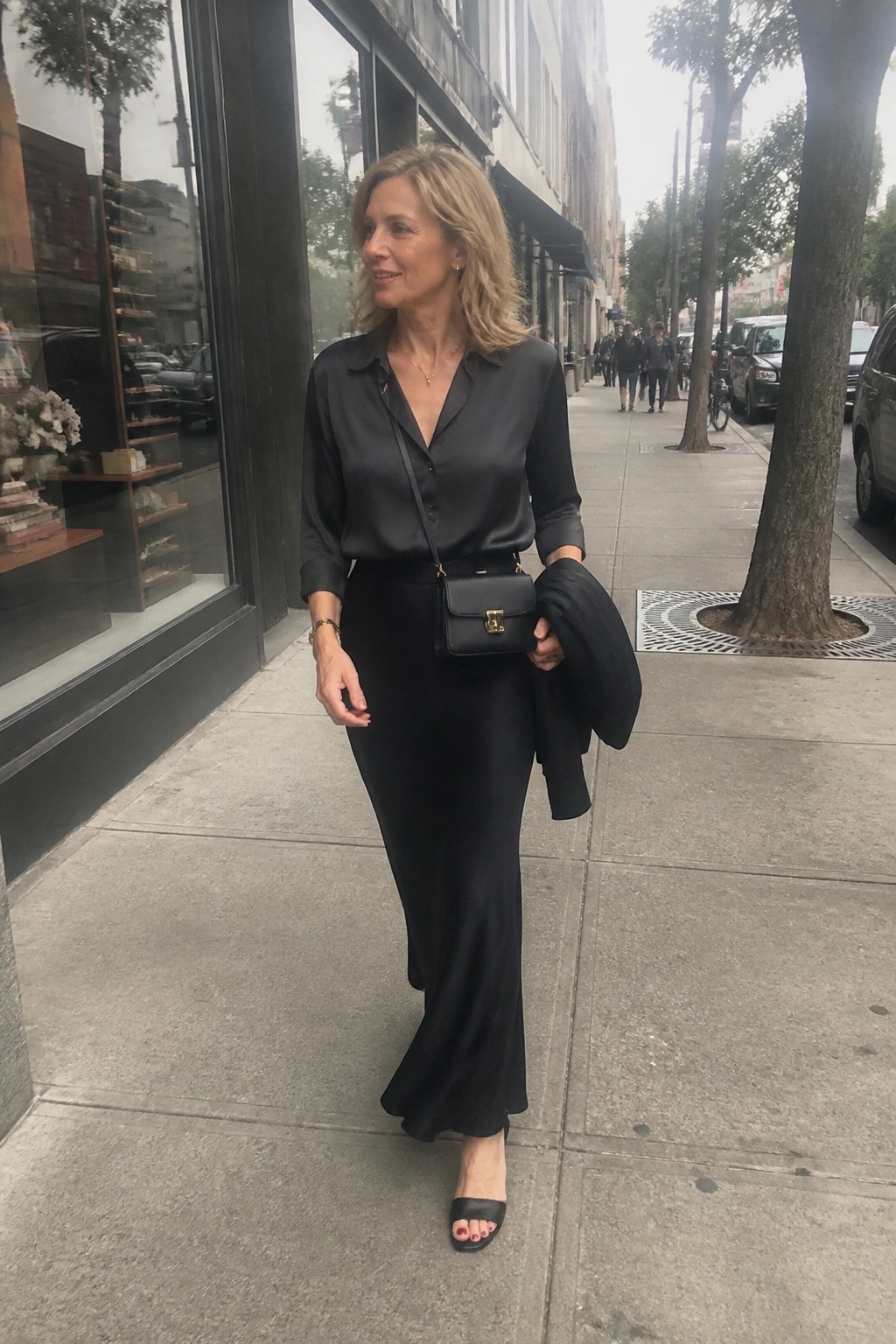Blonde woman in black satin blouse and long skirt walks city sidewalk with handbag.