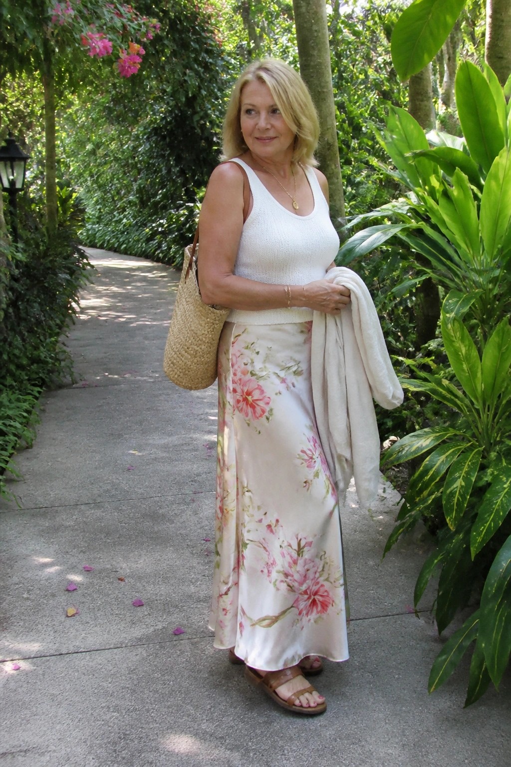 Blonde woman in white top and pink floral skirt holding straw bag on garden path