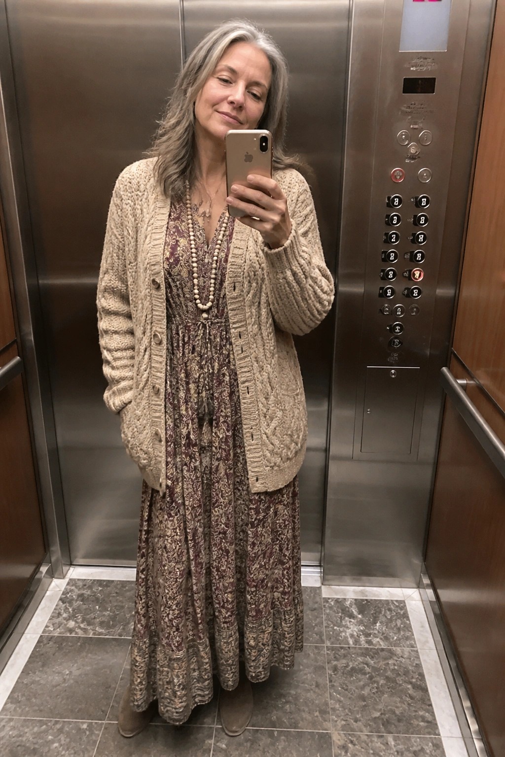 Gray-haired woman in beige cardigan over patterned dress taking selfie in elevator