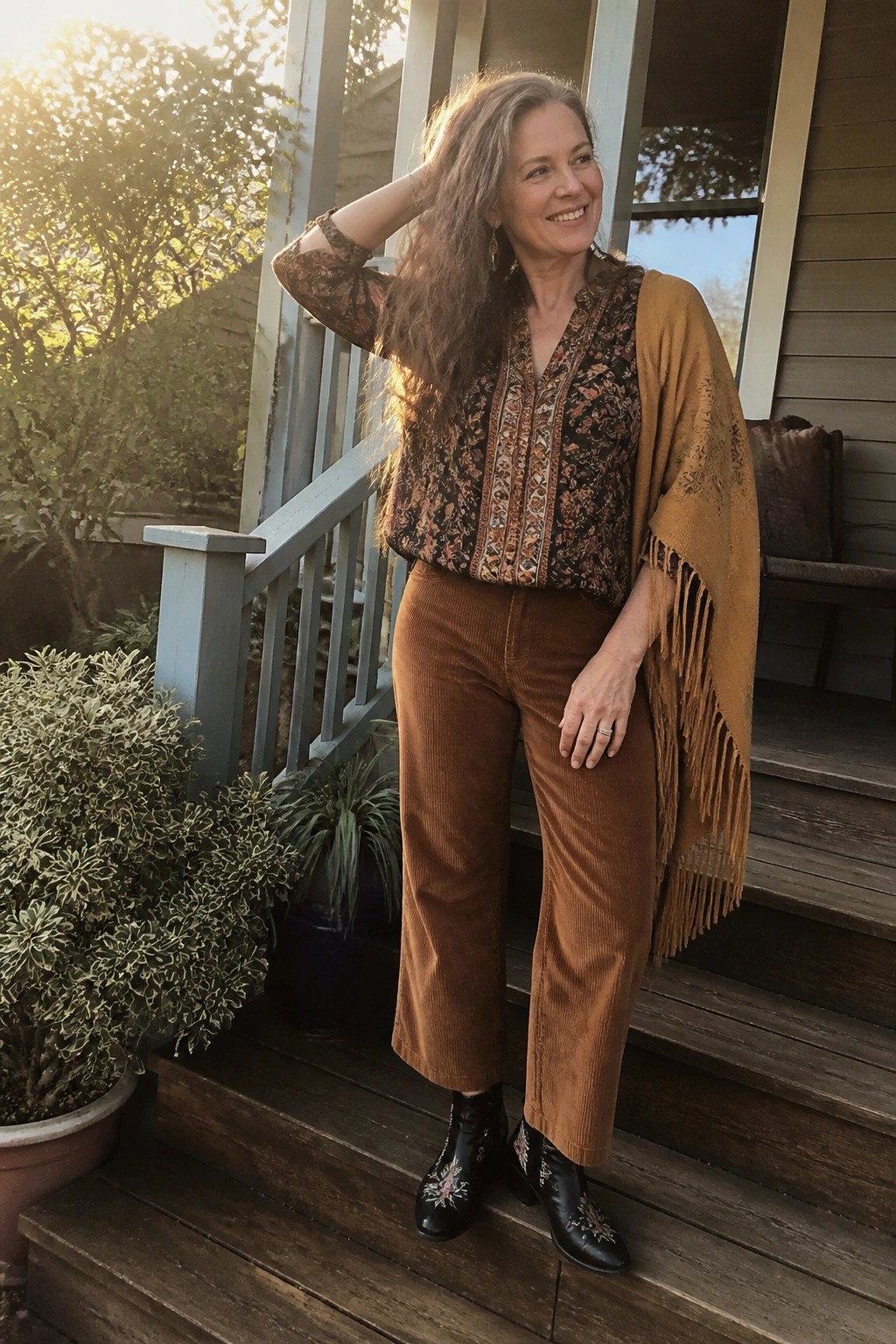 Woman over 50 in brown corduroy wide-leg pants, black floral print blouse, mustard fringed shawl, and black embroidered ankle boots.