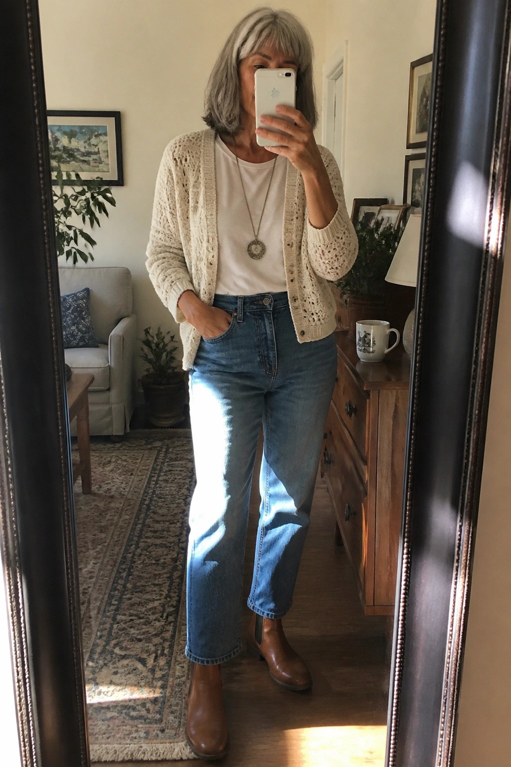Gray-haired woman in cream cardigan and blue jeans taking mirror selfie indoors