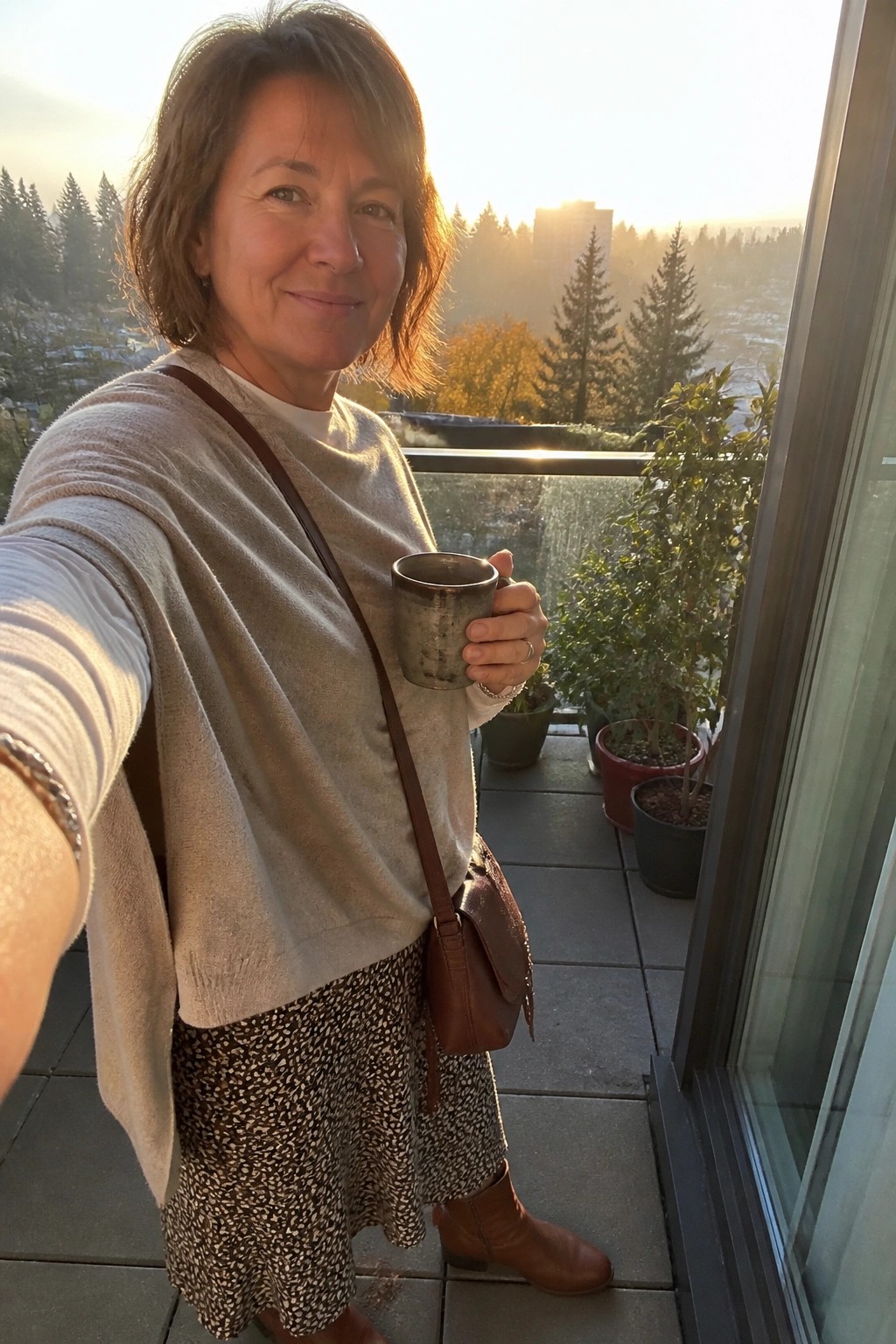 Woman in beige sweater and patterned skirt holding mug on sunny balcony at sunset