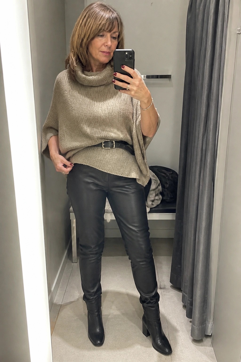 Woman in beige cowl-neck poncho sweater and black leather pants taking mirror selfie