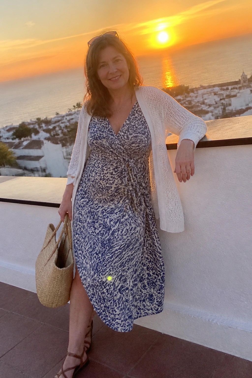 Woman in blue patterned dress and white cardigan holds straw bag at ocean sunset