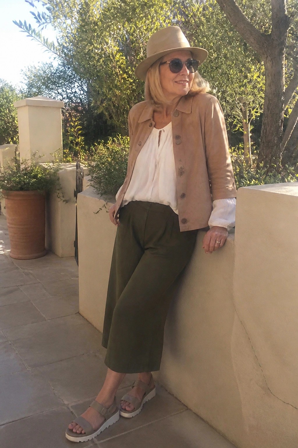 Woman over 50 wearing a tan suede jacket over a white blouse with olive wide leg pants and beige platform sandals plus a matching hat.