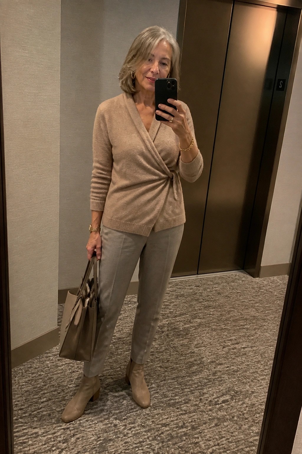 Gray-haired woman in beige wrap sweater and gray pants taking mirror selfie with handbag.