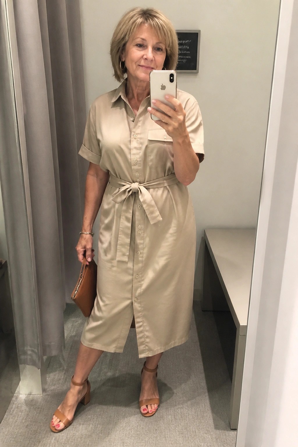 Woman in beige button-up shirt dress taking mirror selfie holding brown clutch