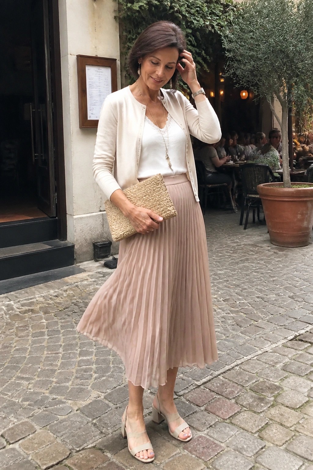 Woman in beige cardigan and pleated skirt holding woven clutch on cobblestones