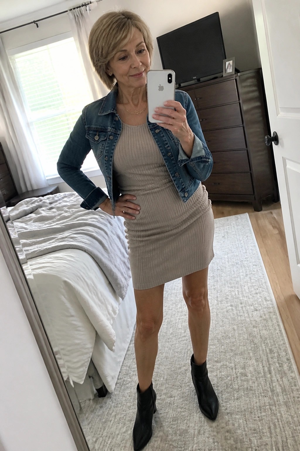 Woman over 50 wearing a beige ribbed mini dress under an open blue denim jacket with black pointed ankle boots and a thin gold necklace while holding a phone for a mirror selfie.