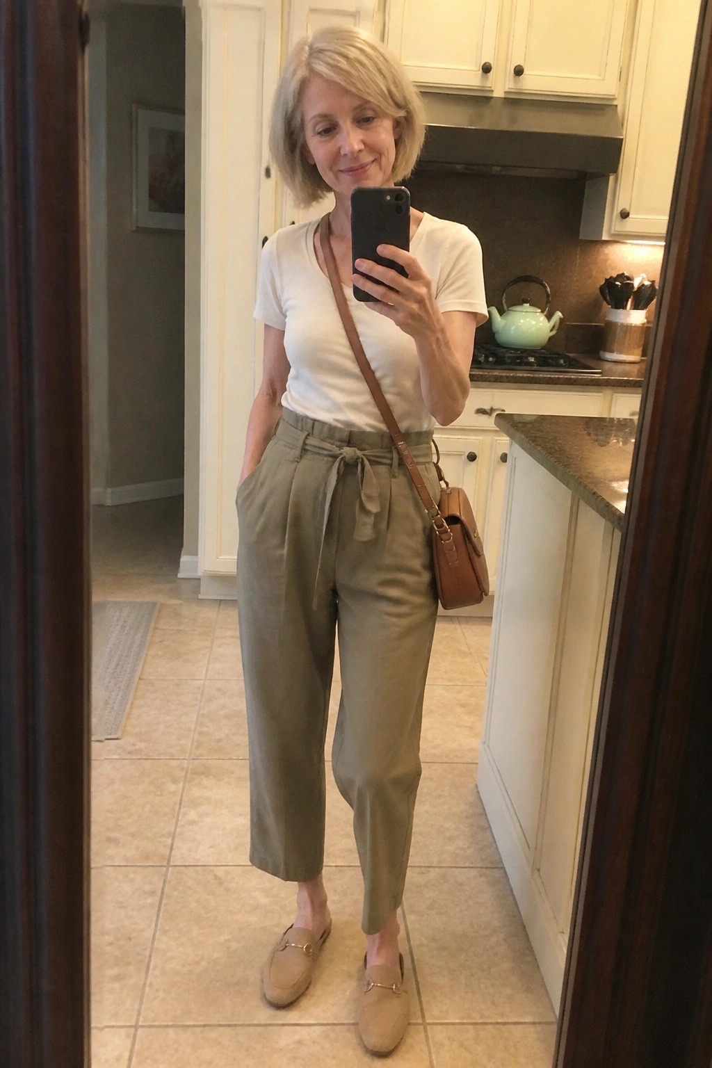 Woman in white t-shirt and olive pants taking mirror selfie in kitchen