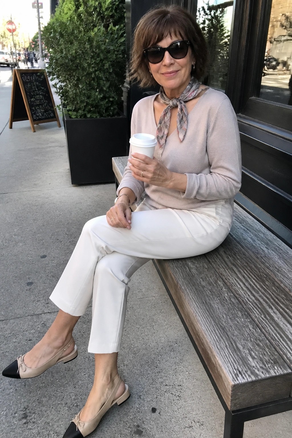 Woman in beige sweater, white pants, and scarf holding coffee on bench
