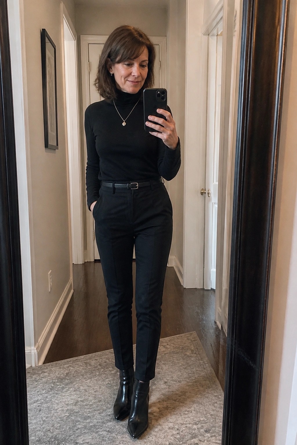 Woman in black turtleneck and pants taking mirror selfie in hallway