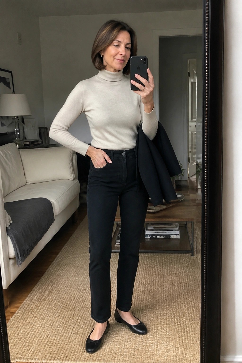 Woman in beige turtleneck, black jeans, and flats taking mirror selfie with phone and jacket