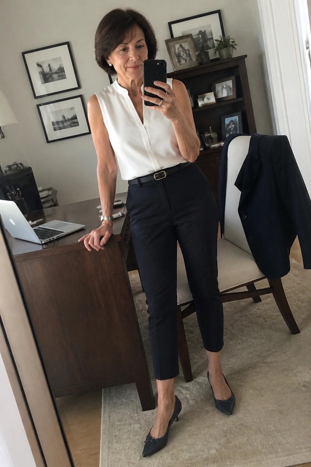 Woman in white sleeveless blouse and navy pants taking mirror selfie in office.