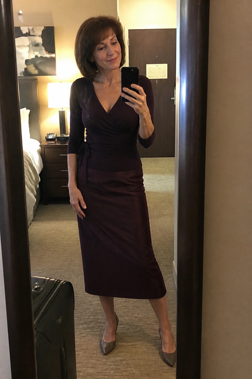 Woman in purple wrap dress taking mirror selfie in hotel room with suitcase
