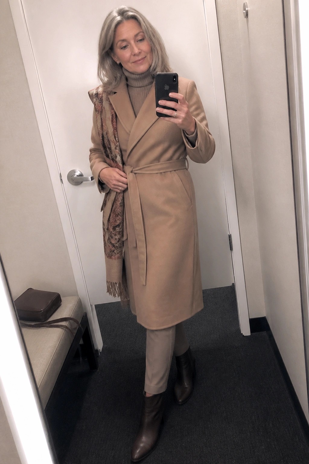 Gray-haired woman in tan coat with patterned scarf taking mirror selfie.