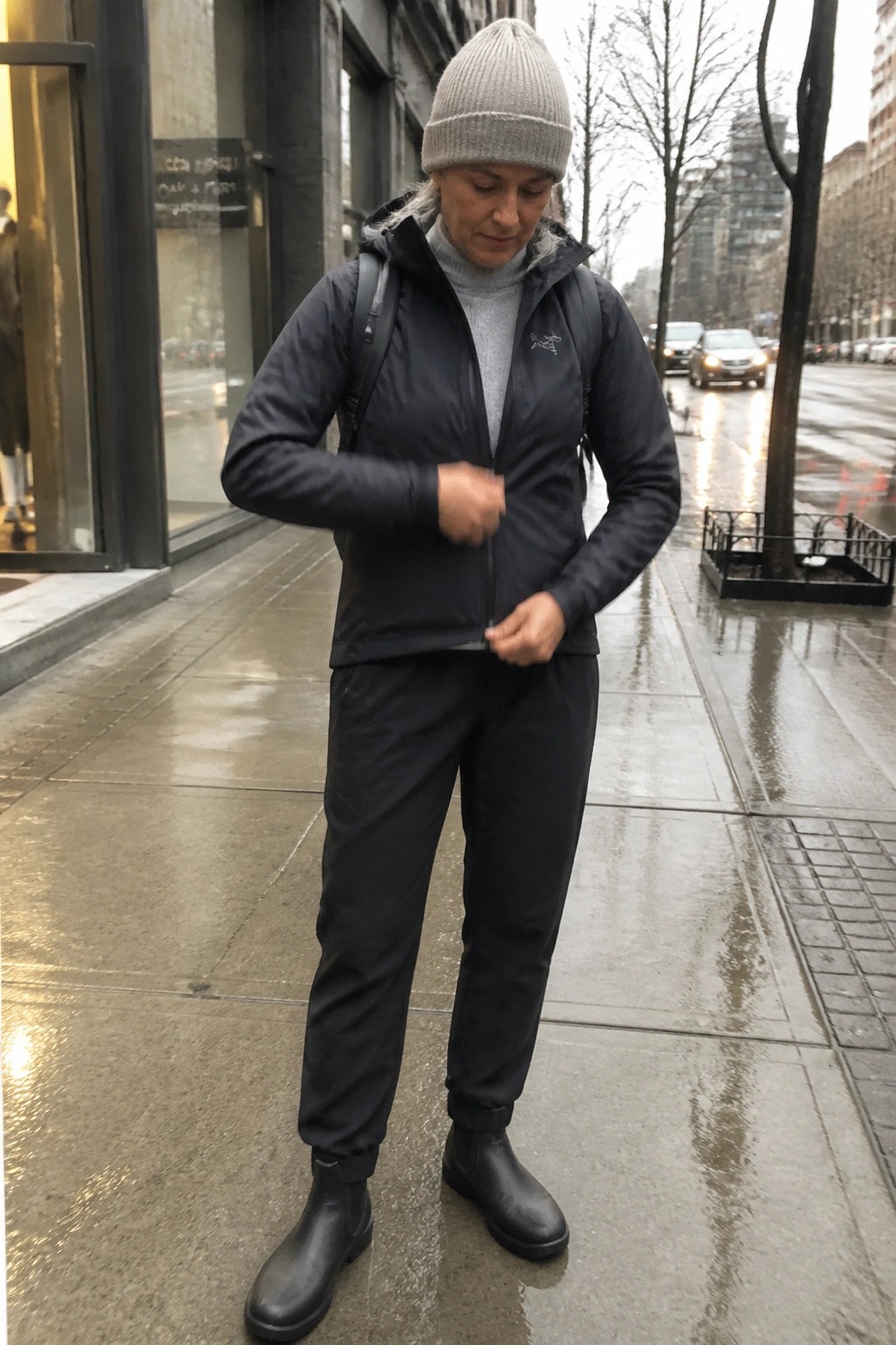 Woman in gray beanie and black jacket zipping up on wet city sidewalk