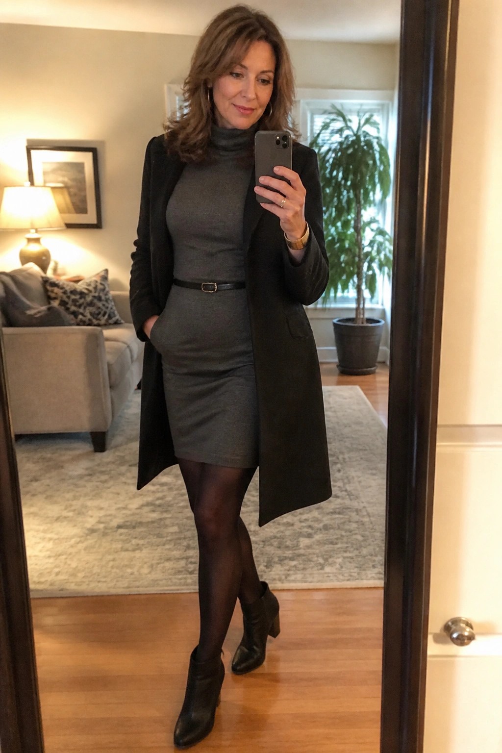 Woman in gray turtleneck dress and black coat taking mirror selfie in living room