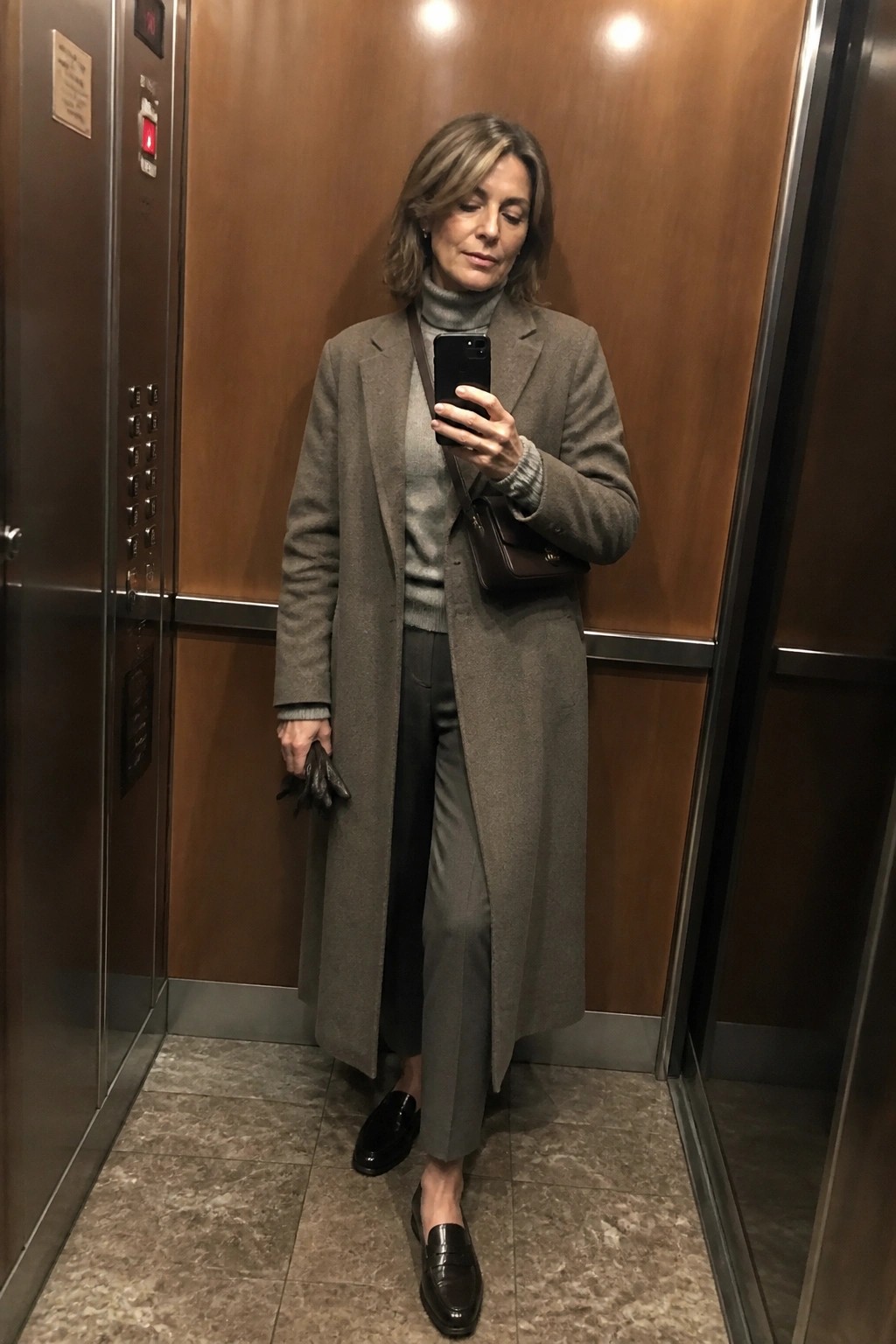 Woman in gray coat and turtleneck holding phone in elevator