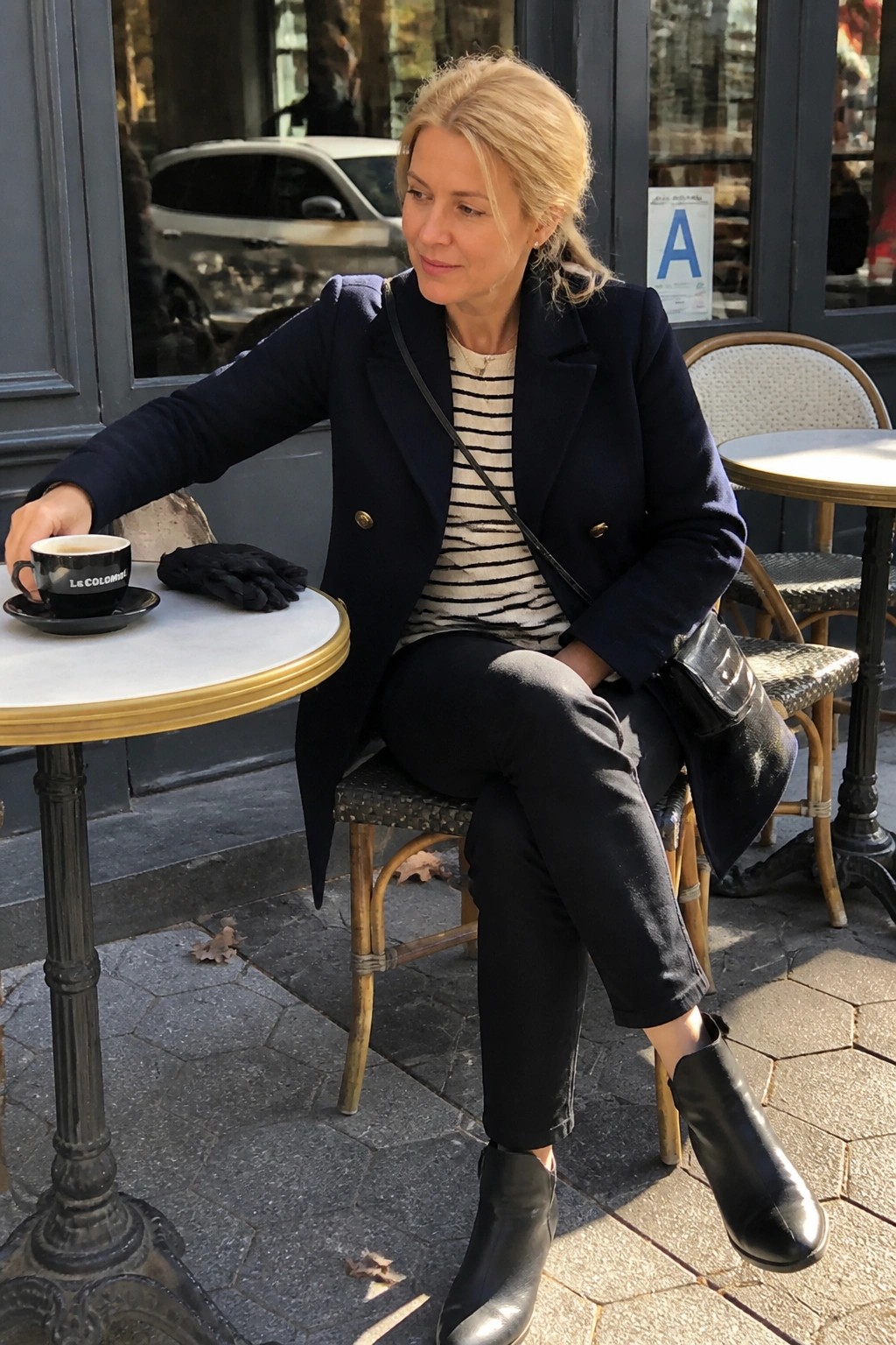 Blonde woman in navy blazer and striped shirt at outdoor café with coffee cup.