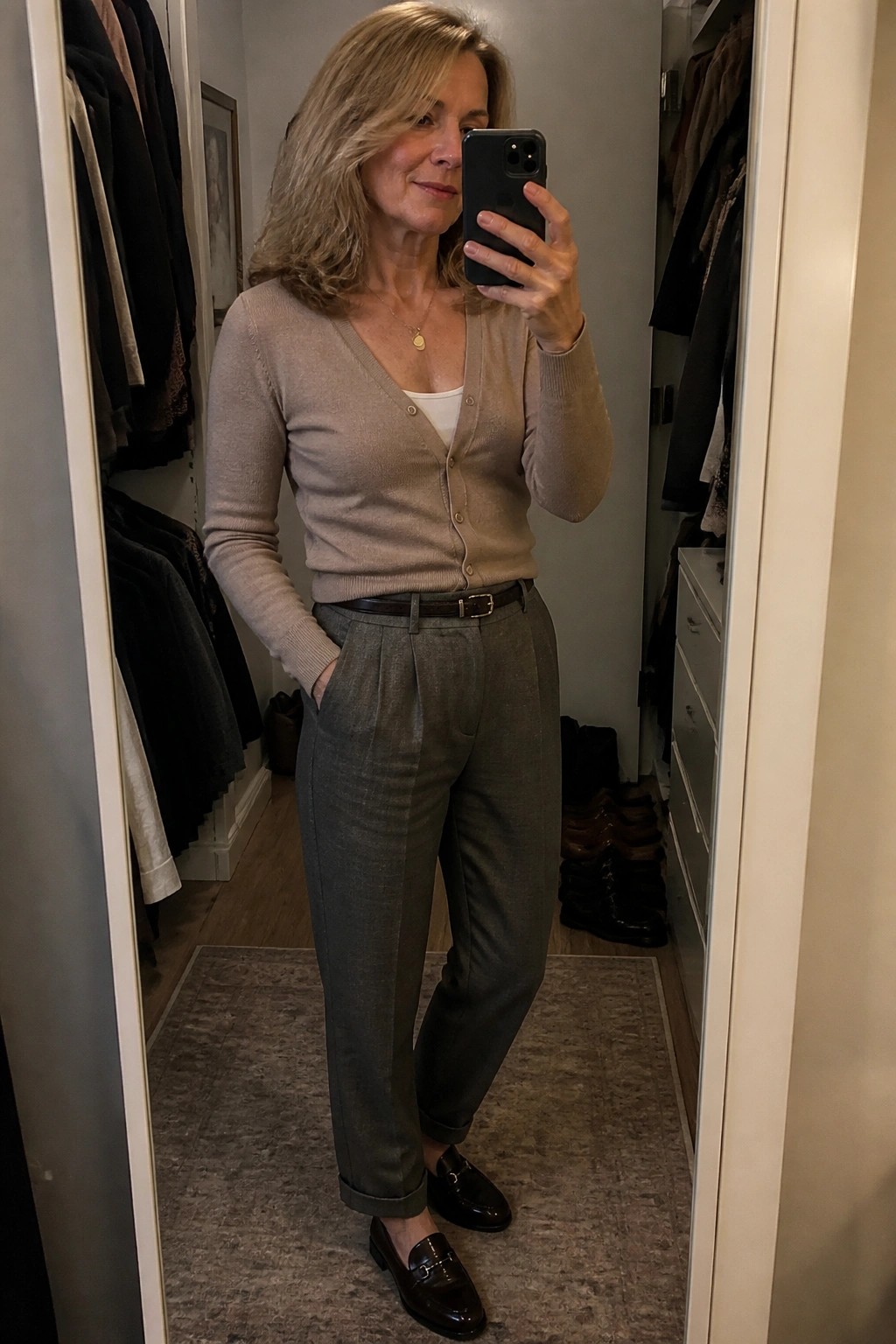 Woman in beige cardigan and gray trousers taking mirror selfie in walk-in closet
