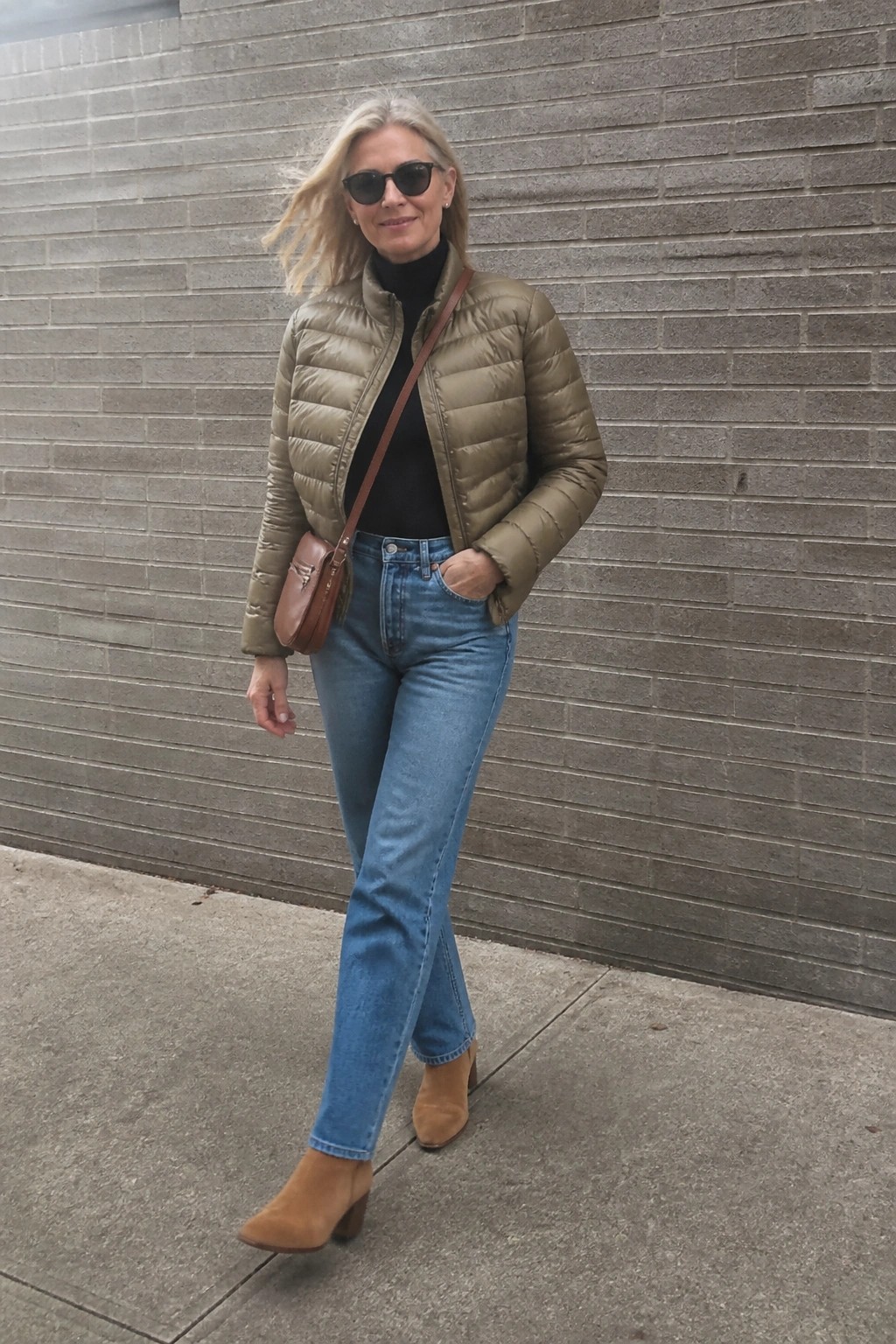 Blonde woman in olive puffer jacket and blue jeans by gray brick wall