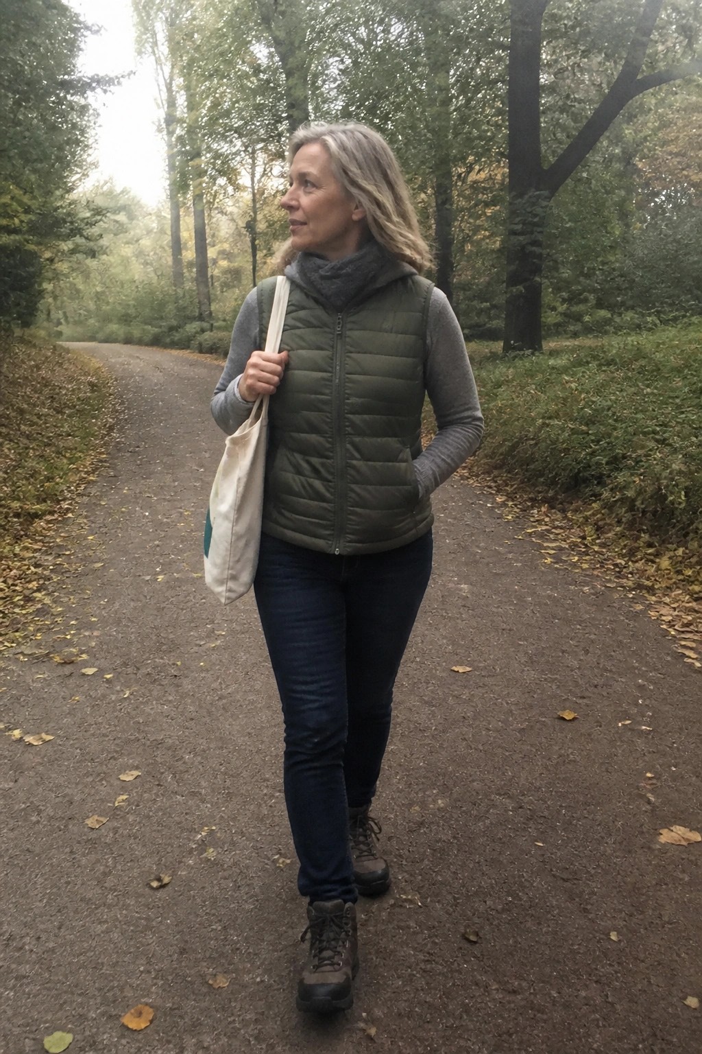 Gray-haired woman in green vest walks autumn forest path with tote bag