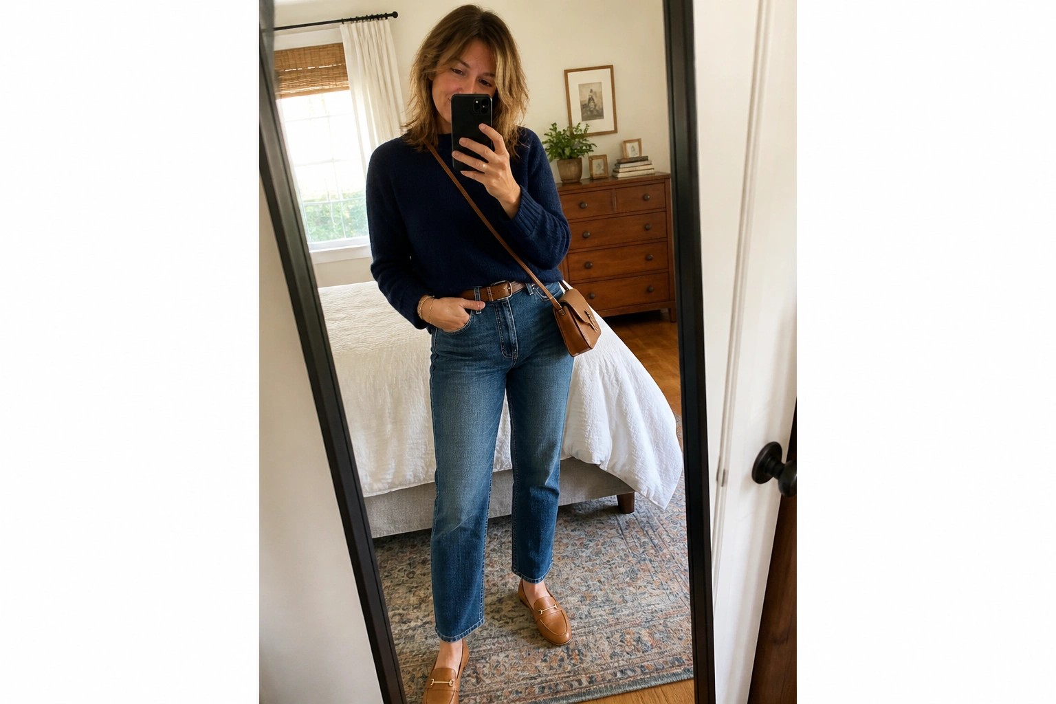Woman in navy sweater and jeans taking mirror selfie in bedroom